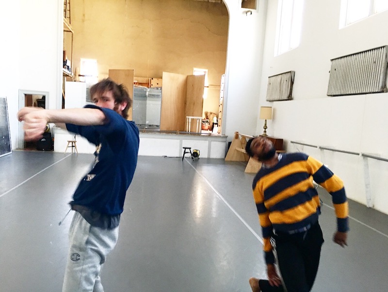 Two men are in mid motion towards the camera. The photo is artfully blurry. They wear rehearsal clothes and are in a sun-filled white studio space.